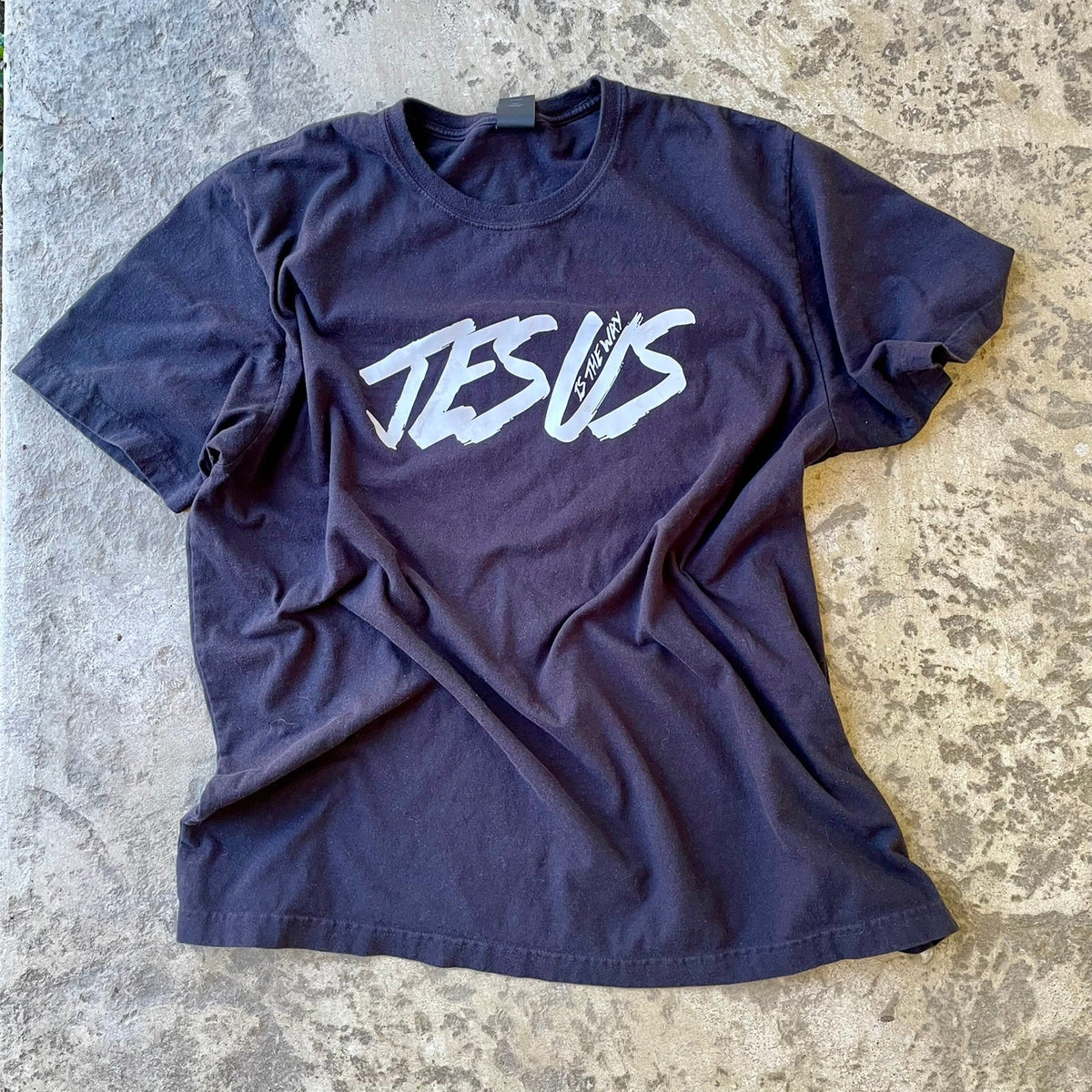 Black t-shirt with a stylized white graphic reading 'JESUS' across the chest, laid flat on a textured concrete surface.