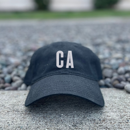 A black baseball cap with 'CA' embroidered in white on the front, resting on a concrete surface with gravel and a blurred building in the background.