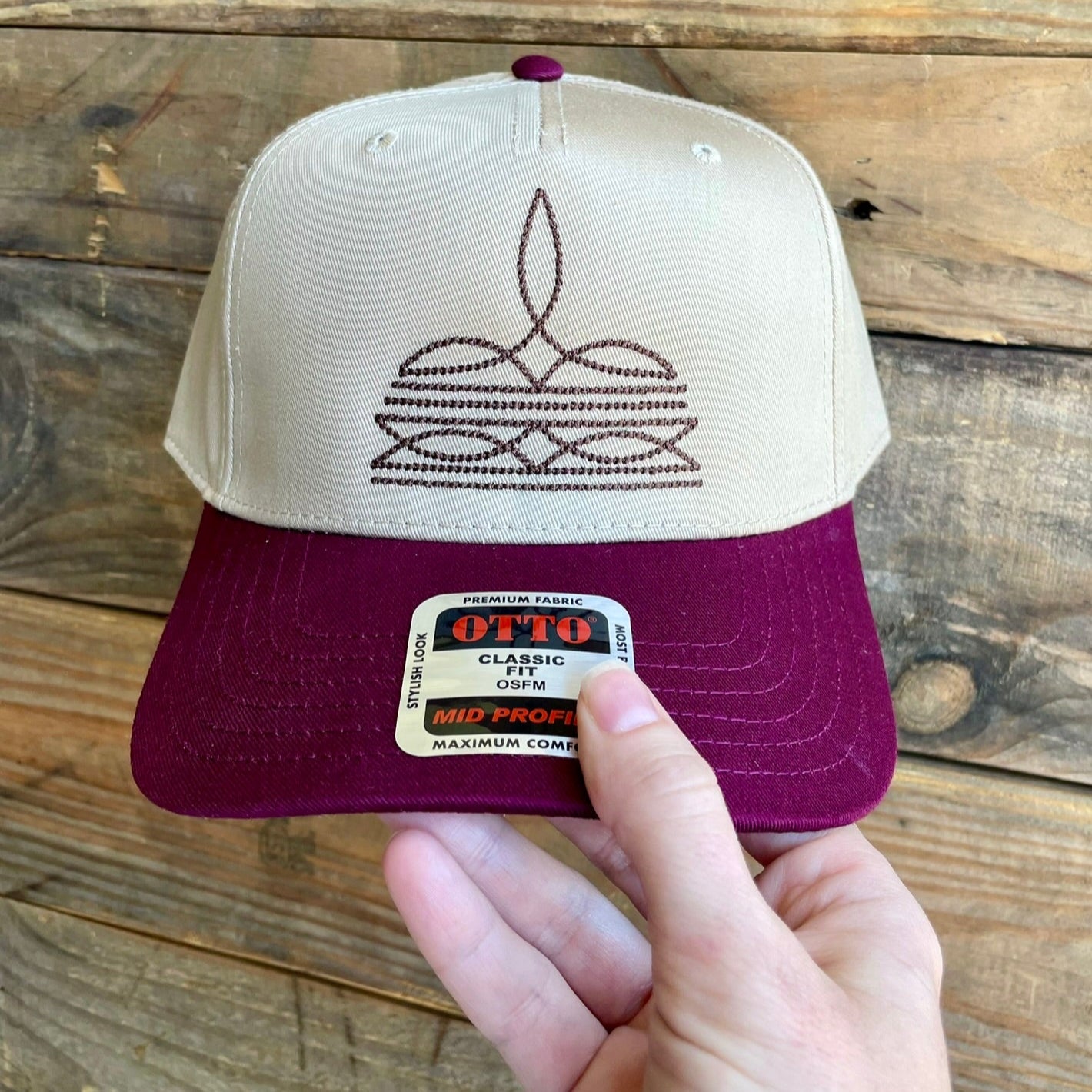 Beige and maroon cap with embroidered boot stitch design held by a hand against a wooden background