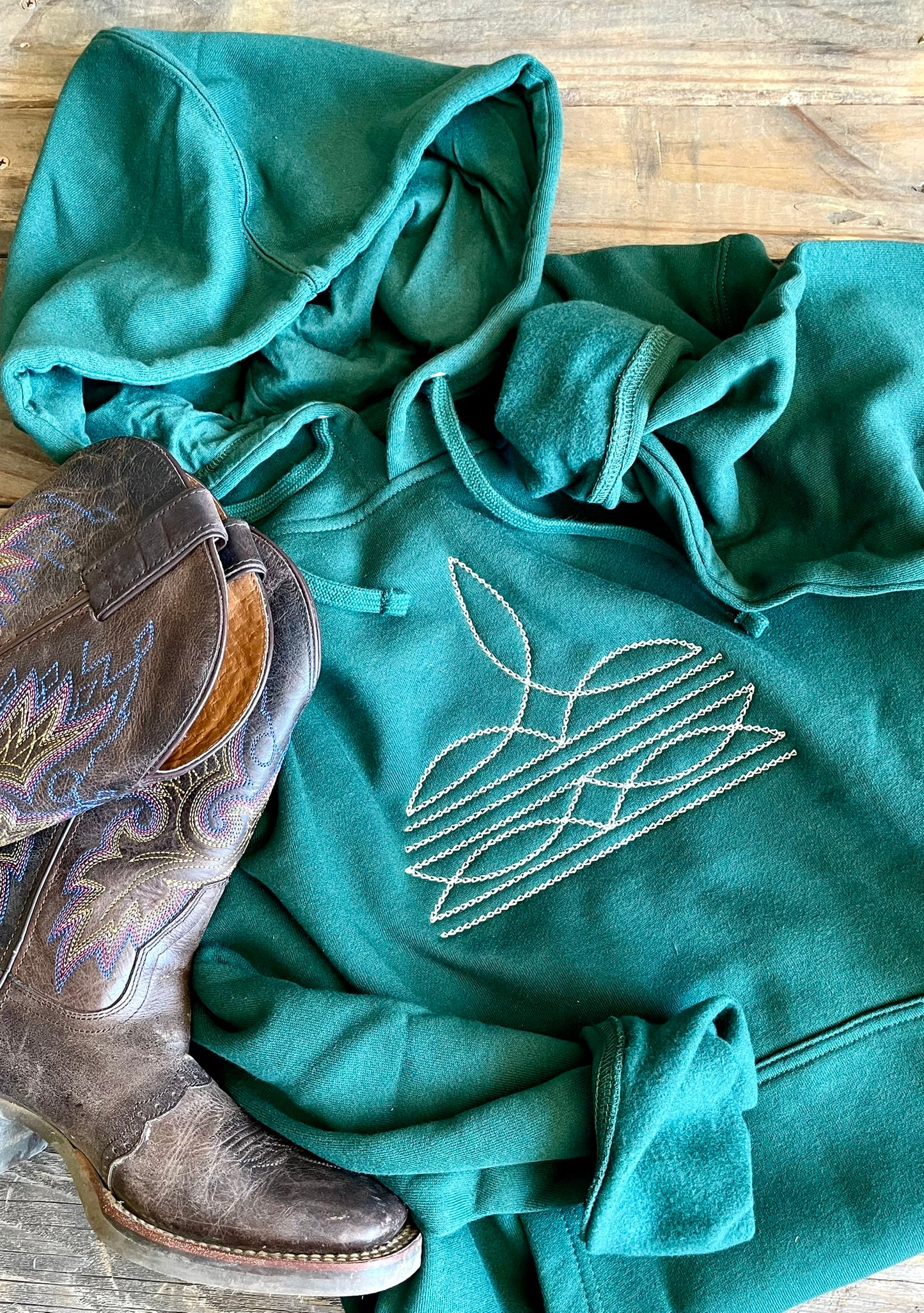 Green hoodie with embroidered design and brown cowboy boot on a wooden surface
