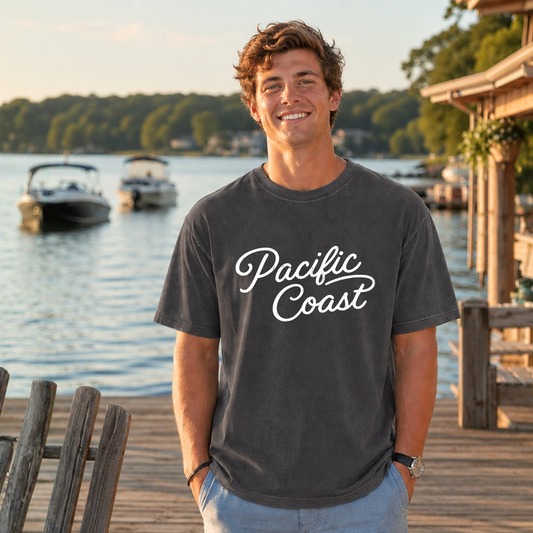 A smiling man wearing a dark charcoal gray t-shirt with 'Pacific Coast' printed in white script across the chest, standing on a wooden dock with a lake and boats in the background.