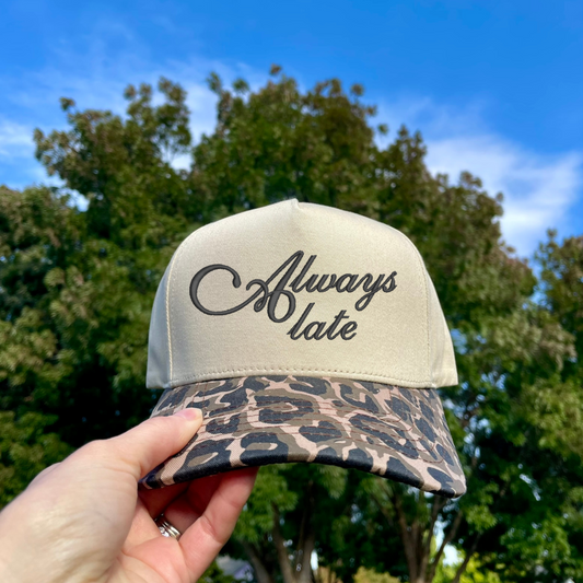 Beige cap with leopard print brim and 'Always late' text held against a blue sky with trees.