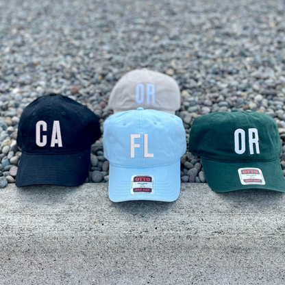 Three baseball caps lined up on a concrete ledge outdoors: a black cap with white 'CA' lettering, a light blue cap with white 'FL' lettering, and a dark green cap with white 'OR' lettering. A light gray cap with pale blue 'OR' lettering sits behind them on gravel.