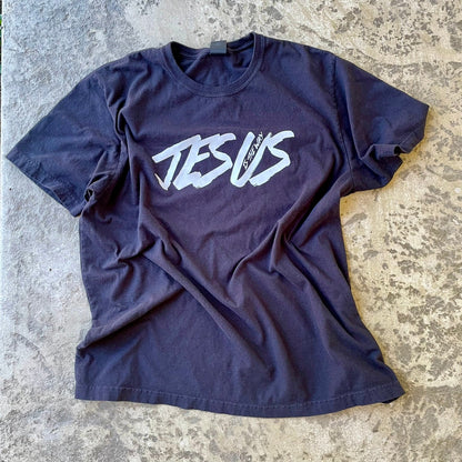 Black t-shirt with a stylized white graphic reading 'JESUS' across the chest, laid flat on a textured concrete surface.
