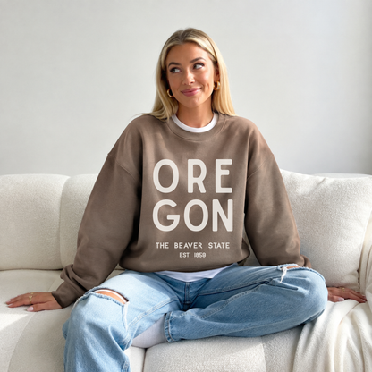 A woman sitting cross-legged on a white couch wearing a taupe brown crewneck sweatshirt with 'OREGON THE BEAVER STATE EST. 1969' printed in white text, paired with light blue ripped jeans.