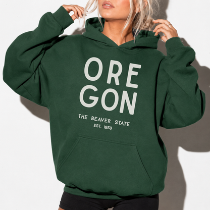 A woman wearing a dark green pullover hoodie with 'OREGON THE BEAVER STATE EST. 1869' printed in white on the front, set against a plain white background.