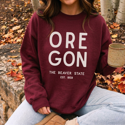 A person wearing a burgundy crewneck sweatshirt with 'OREGON THE BEAVER STATE EST. 1859' printed in white on the front, sitting outdoors on a stone bench surrounded by fallen autumn leaves and holding a ceramic mug.