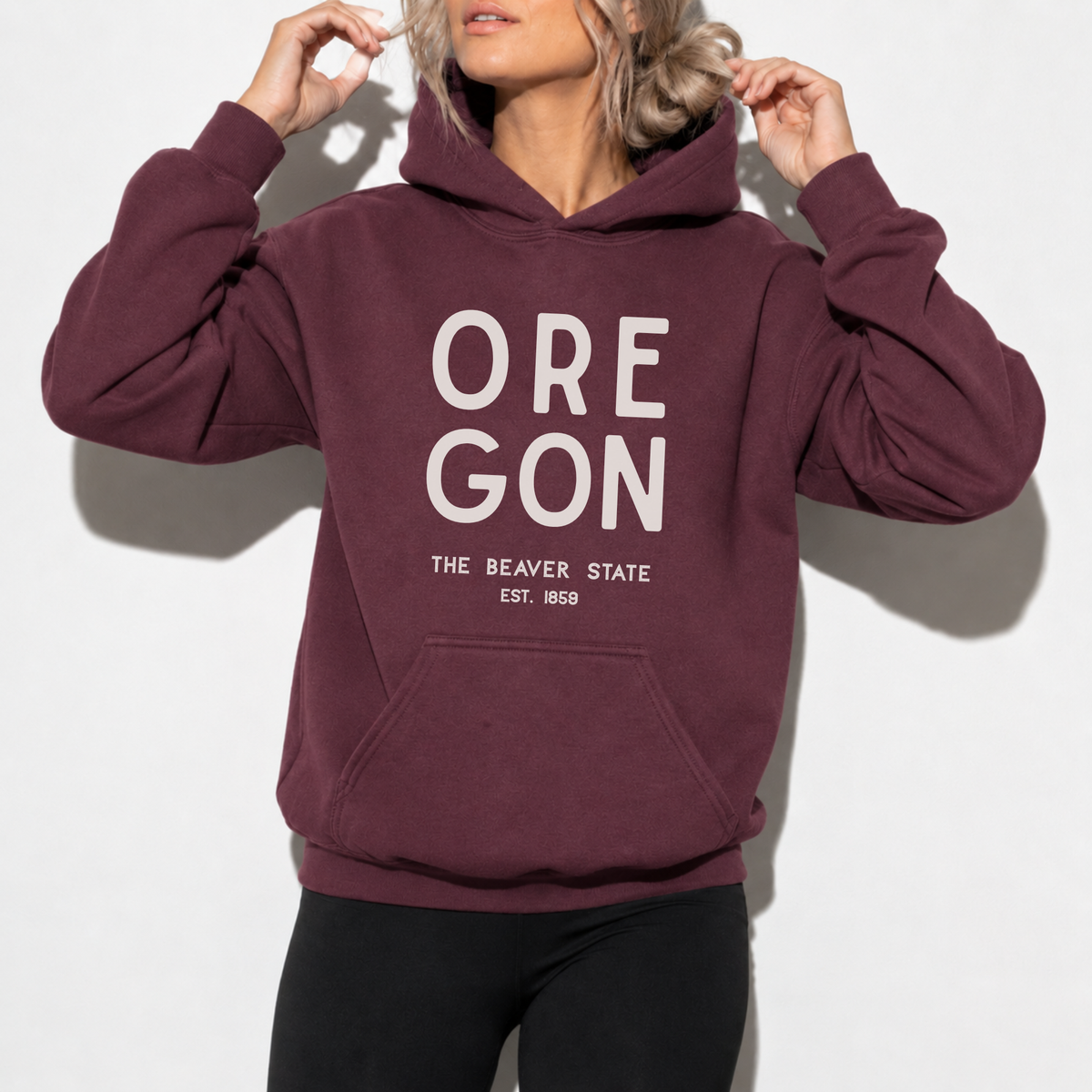 A woman wearing a maroon pullover hoodie with 'OREGON THE BEAVER STATE EST. 1850' printed in white on the front, paired with black leggings, against a plain white background.