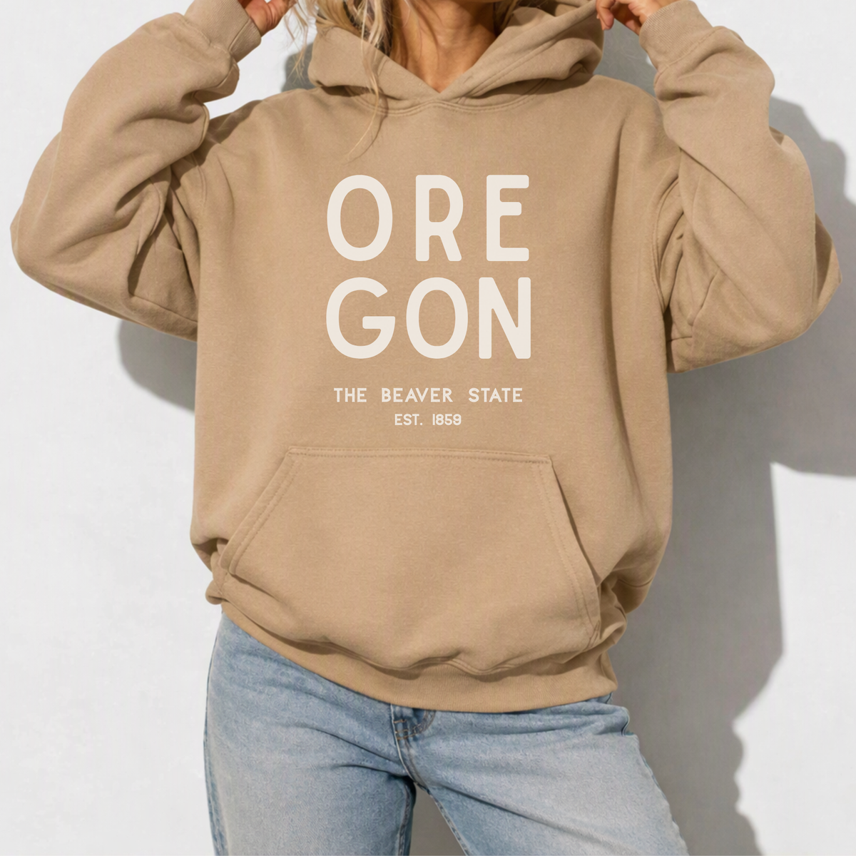 A person wearing a tan pullover hoodie with large white 'OREGON' text and smaller 'THE BEAVER STATE EST. 1858' text on the front, paired with light blue jeans against a light gray wall.