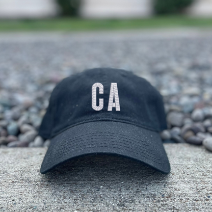 A black baseball cap with 'CA' embroidered in white on the front, resting on a concrete surface with gravel and a blurred building in the background.
