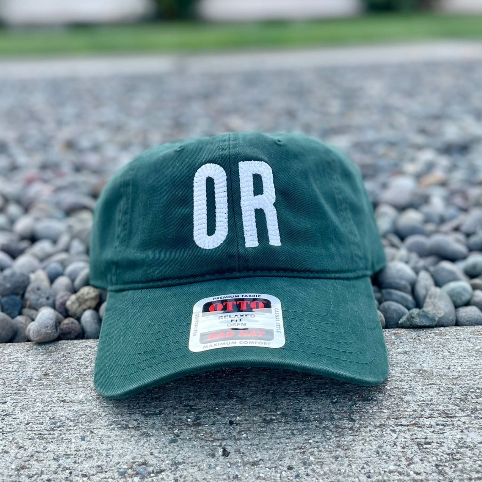 A dark green baseball cap with 'OR' embroidered in white on the front, resting on a concrete ledge with gravel and a blurred house in the background.