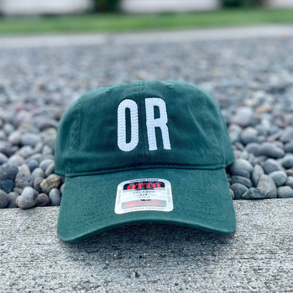 A dark green baseball cap with 'OR' embroidered in white on the front, resting on a concrete ledge with gravel and a blurred house in the background.