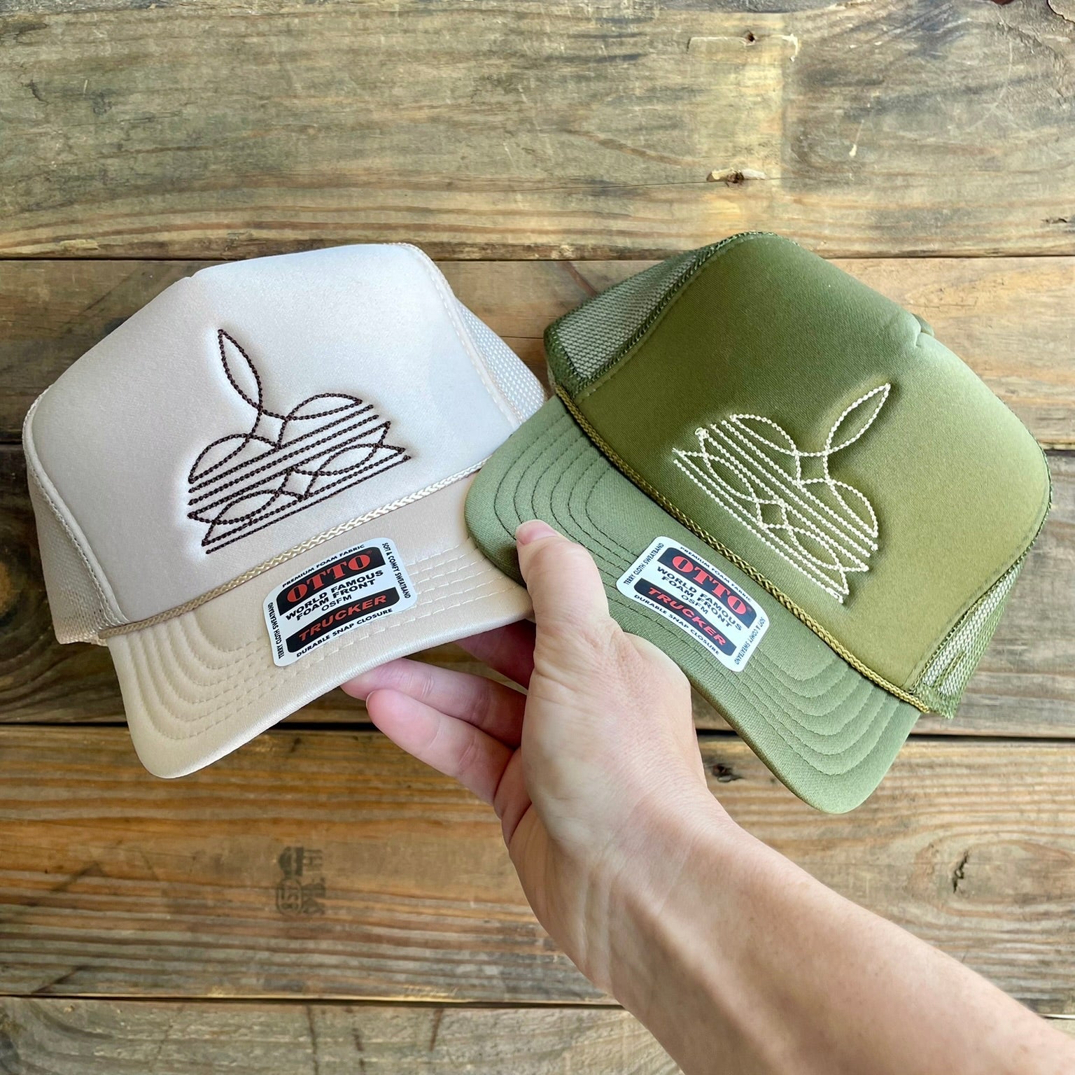 Two embroidered caps, one beige and one green, held by a hand on a wooden surface.