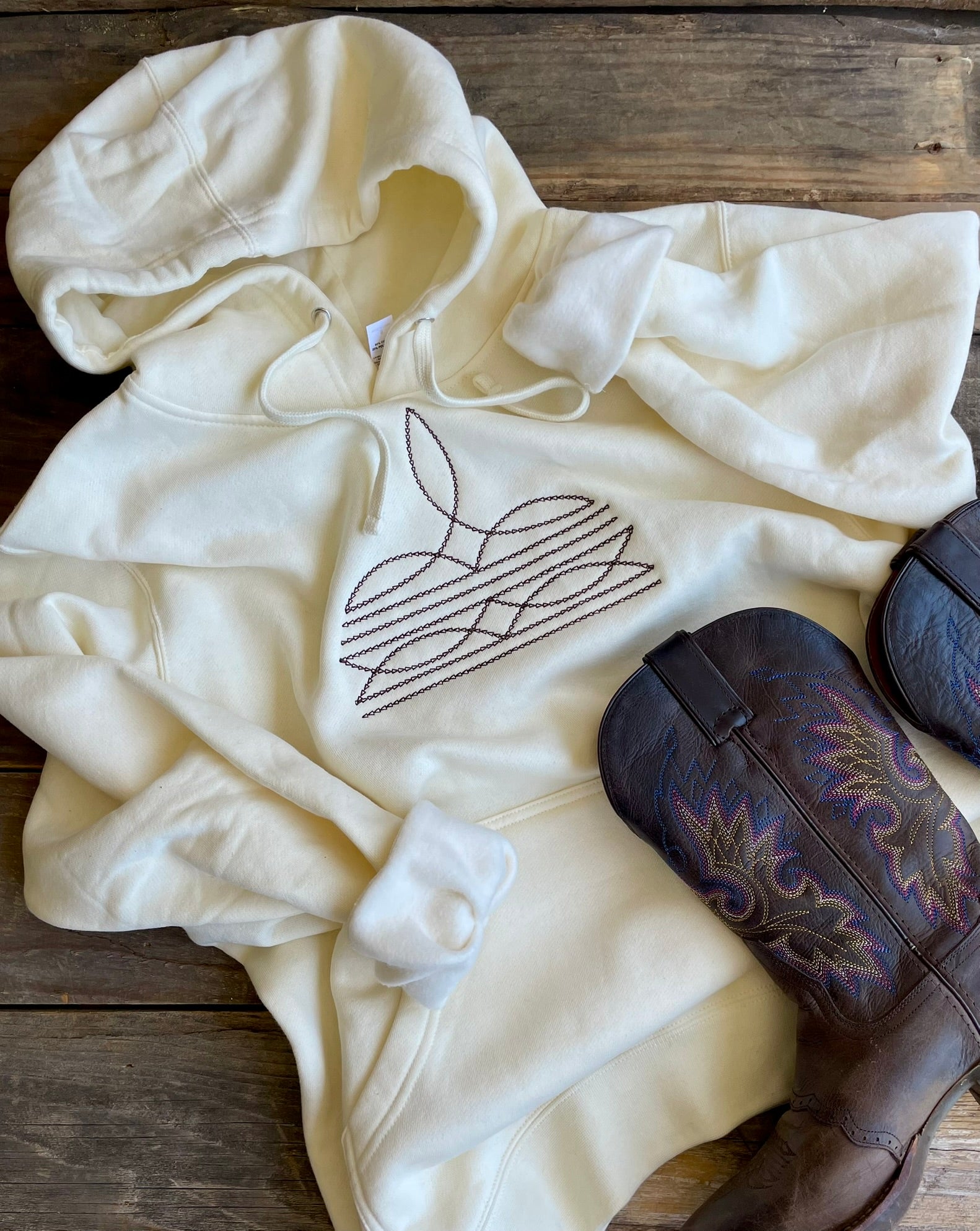 White hoodie with a boot stitch design on a wooden surface with cowboy boots.