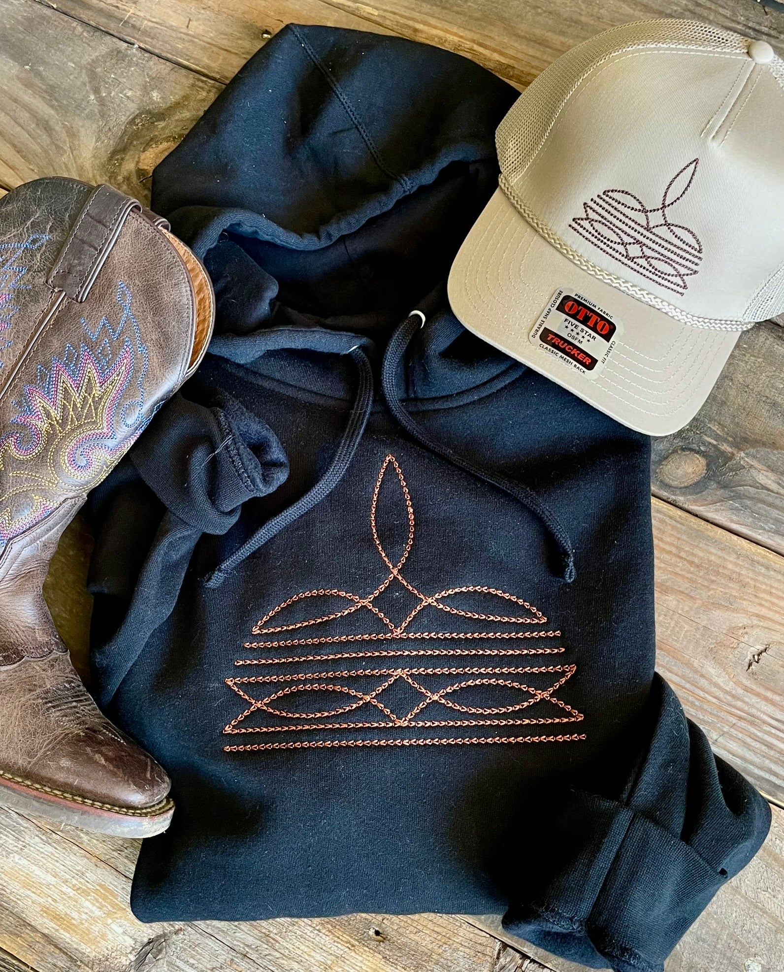 Black hoodie with intricate design, beige cap, and cowboy boots on a wooden surface