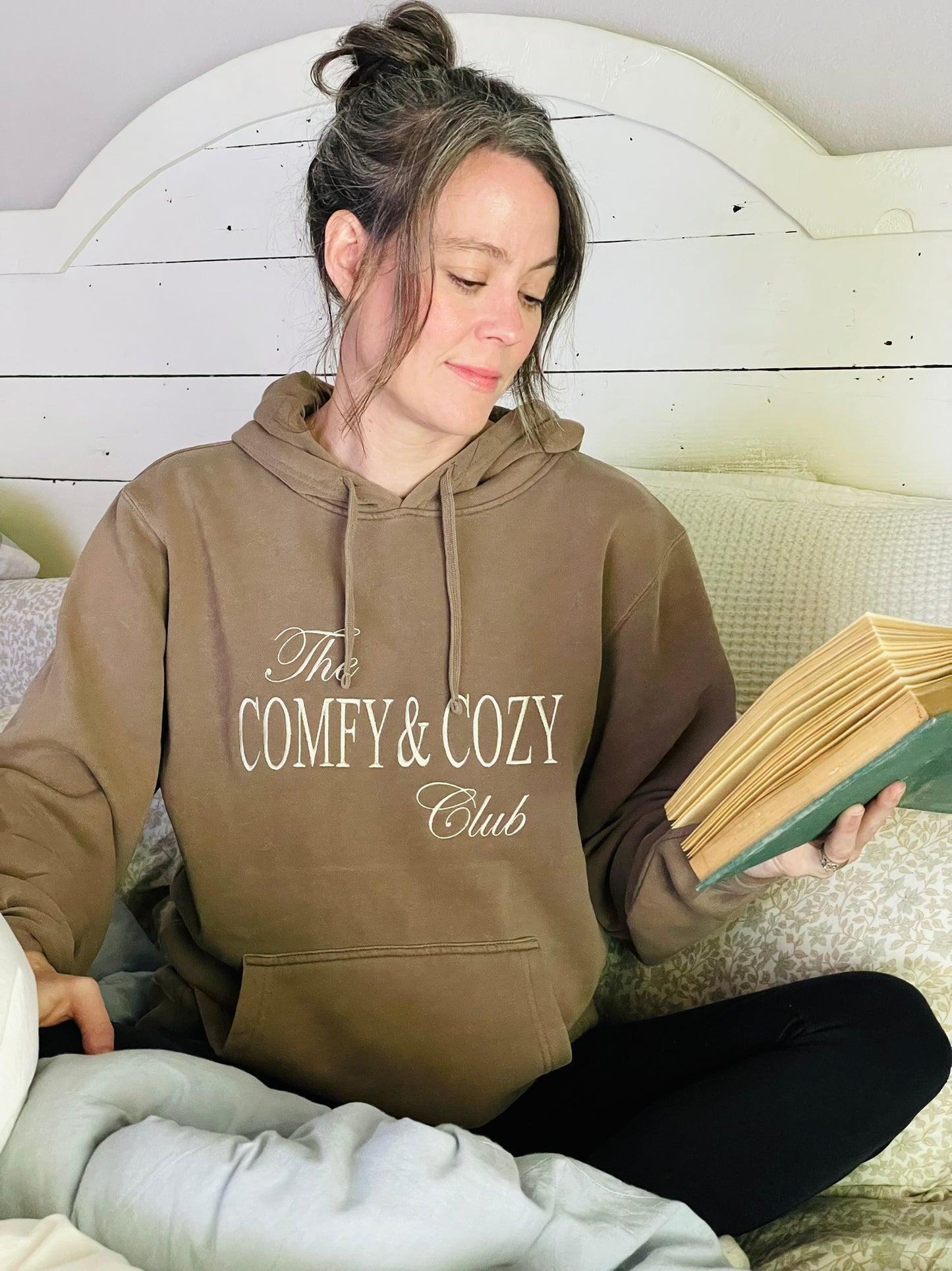 Woman wearing a hoodie with 'The Comfy & Cozy Club' text, reading a book.