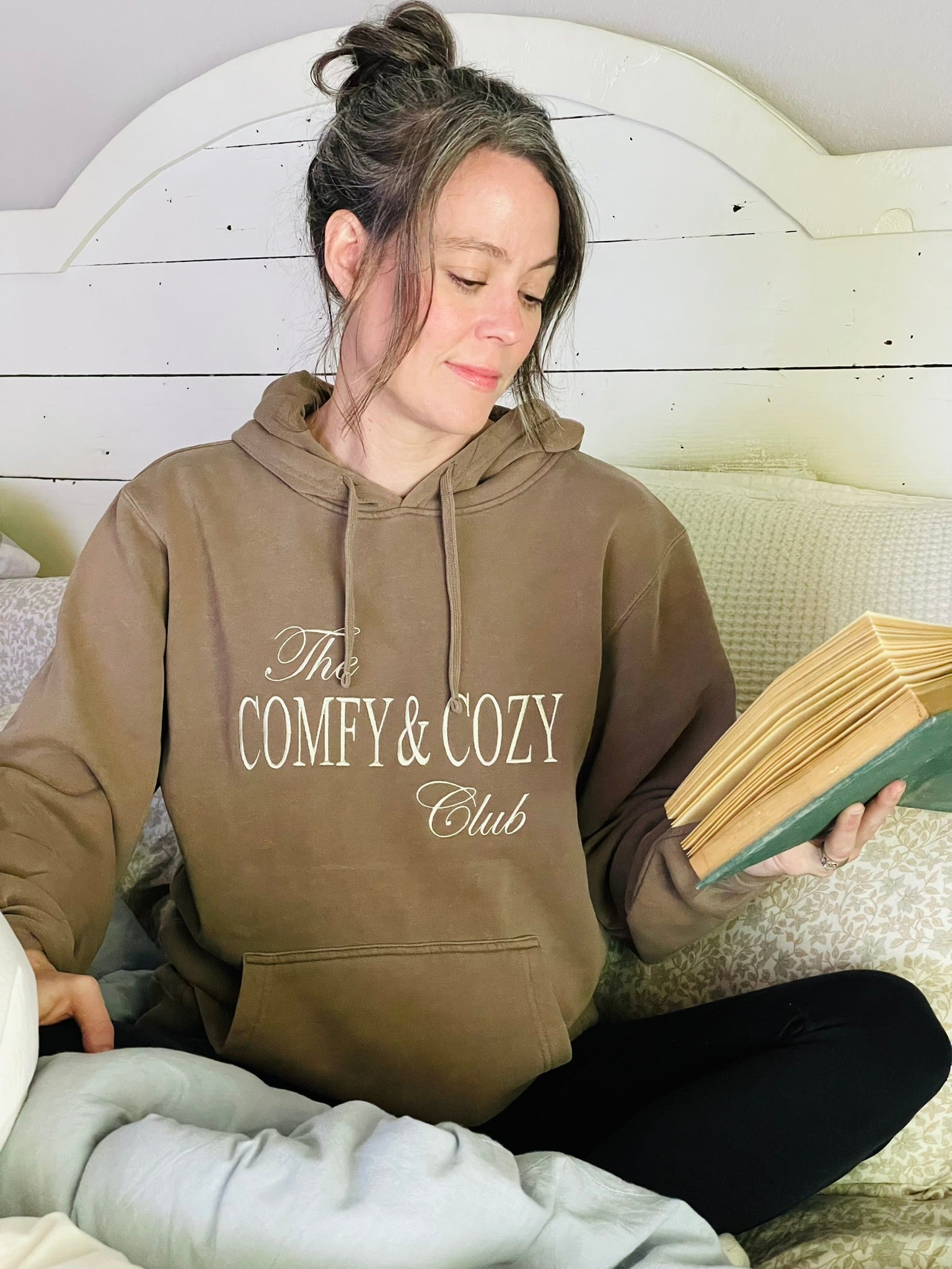 Woman wearing a hoodie with 'The Comfy & Cozy Club' text, reading a book.