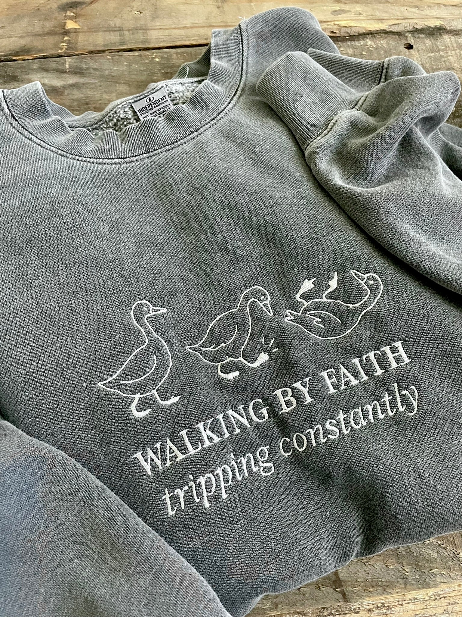 faded black crewneck embroidered with 3 geese and phrase "Walking by Faith tripping constantly"