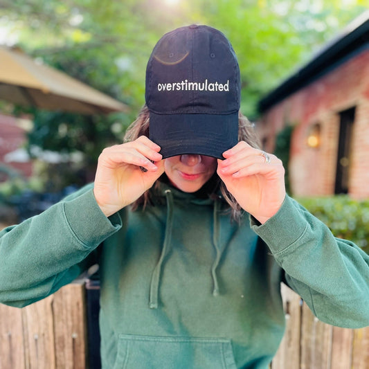 Person wearing a green hoodie and black cap with 'overstimulated' text outdoors.