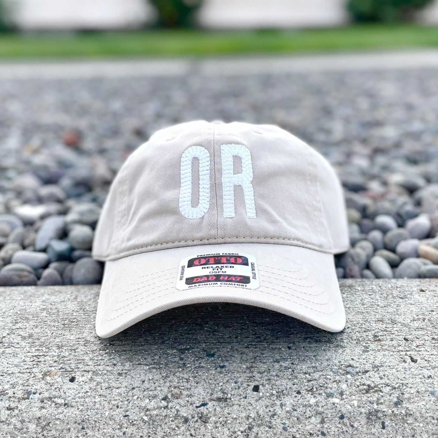 A light beige baseball cap with white embroidered 'OR' lettering on the front, resting on a concrete ledge with gravel and a blurred house in the background.