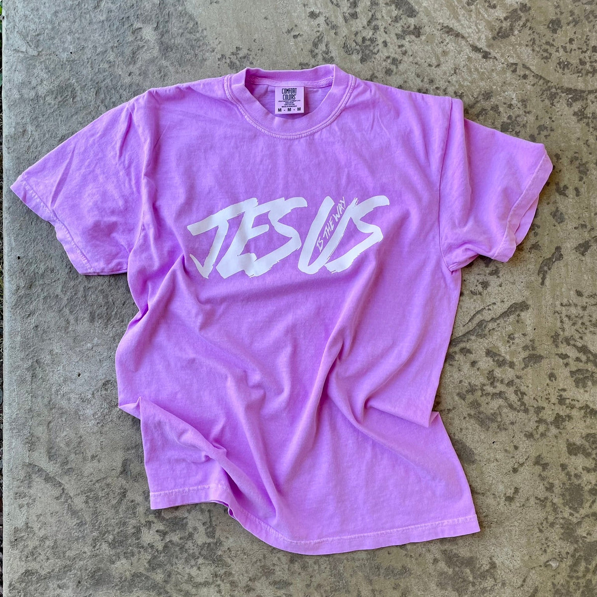 A light purple short-sleeve t-shirt with 'JESUS' printed across the chest in a white brush-stroke font, laid flat on a textured concrete surface.