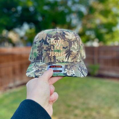 A camouflage baseball cap held up by a hand in an outdoor backyard setting. The front of the cap features the text 'LOVE LIKE JESUS' in a gold embroidered font, and a white sticker is visible on the brim.