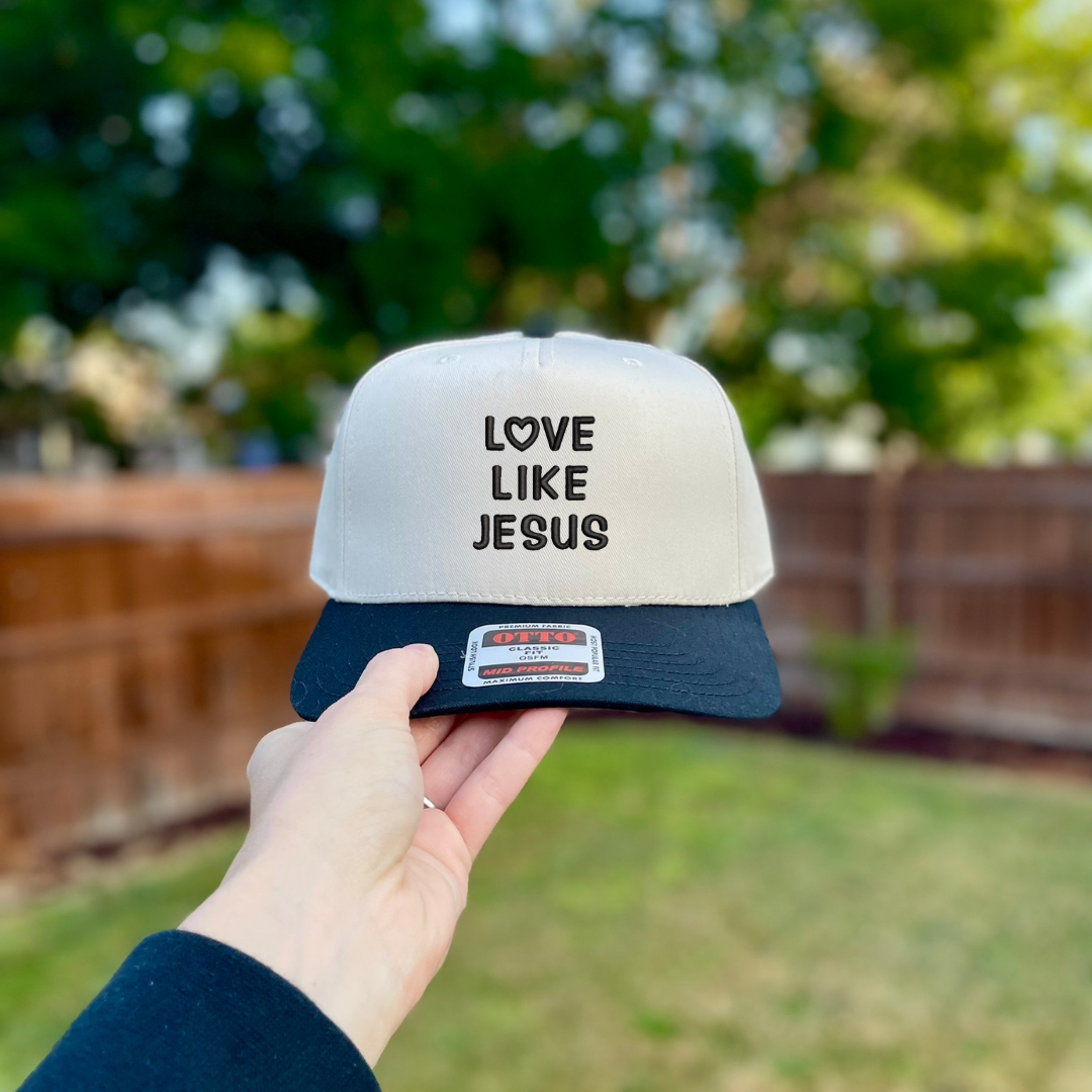 A hand holds a two-tone snapback cap outdoors with a blurred backyard background. The cap has a white crown with black text reading 'LOVE LIKE JESUS' and a dark navy blue brim.