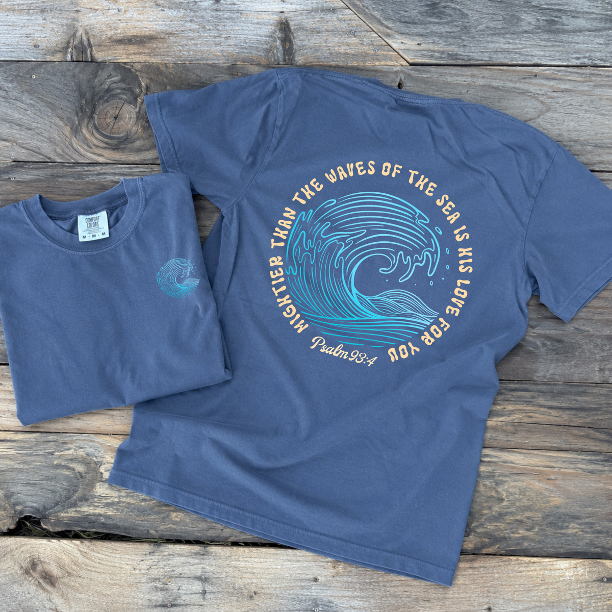 A navy blue short-sleeve t-shirt with a large ocean wave graphic and the text 'MIGHTIER THAN THE WAVES OF THE SEA IS HIS LOVE FOR YOU Psalm 33:4' printed on the back, shown alongside a smaller folded view of the same shirt on a rustic wooden plank background.