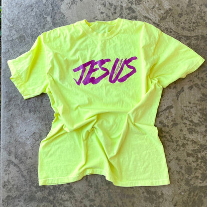 A neon yellow-green t-shirt with the word 'JESUS' printed across the chest in purple script, laid flat on a textured concrete floor.