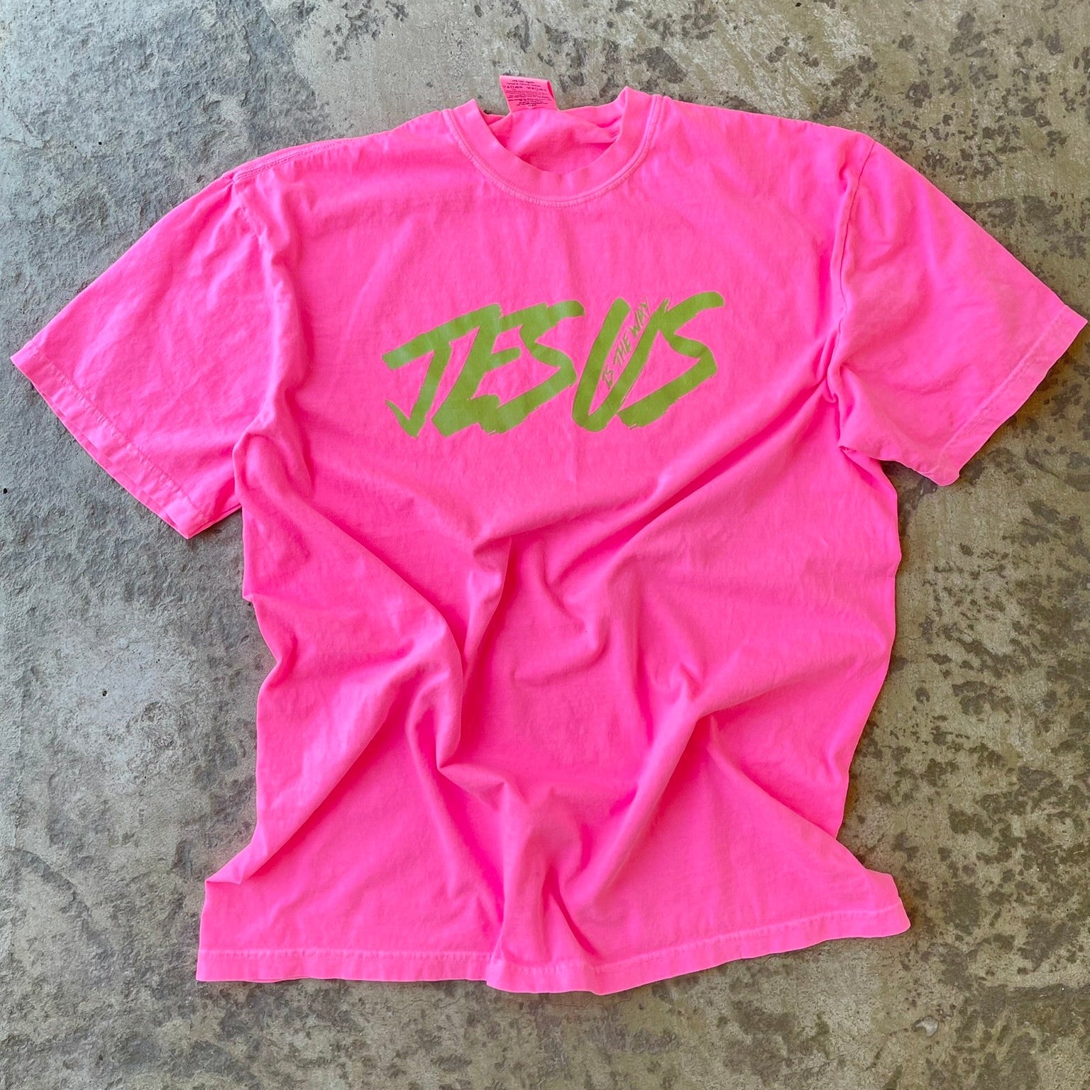 A bright pink short-sleeve t-shirt with the word 'JESUS' printed across the chest in a lime green graffiti-style font, laid flat on a textured gray concrete floor.