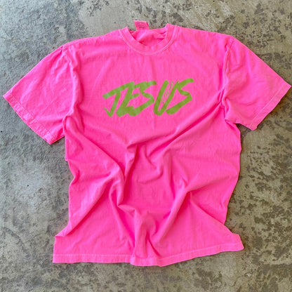 A bright pink short-sleeve t-shirt with the word 'JESUS' printed across the chest in a lime green graffiti-style font, laid flat on a textured gray concrete floor.
