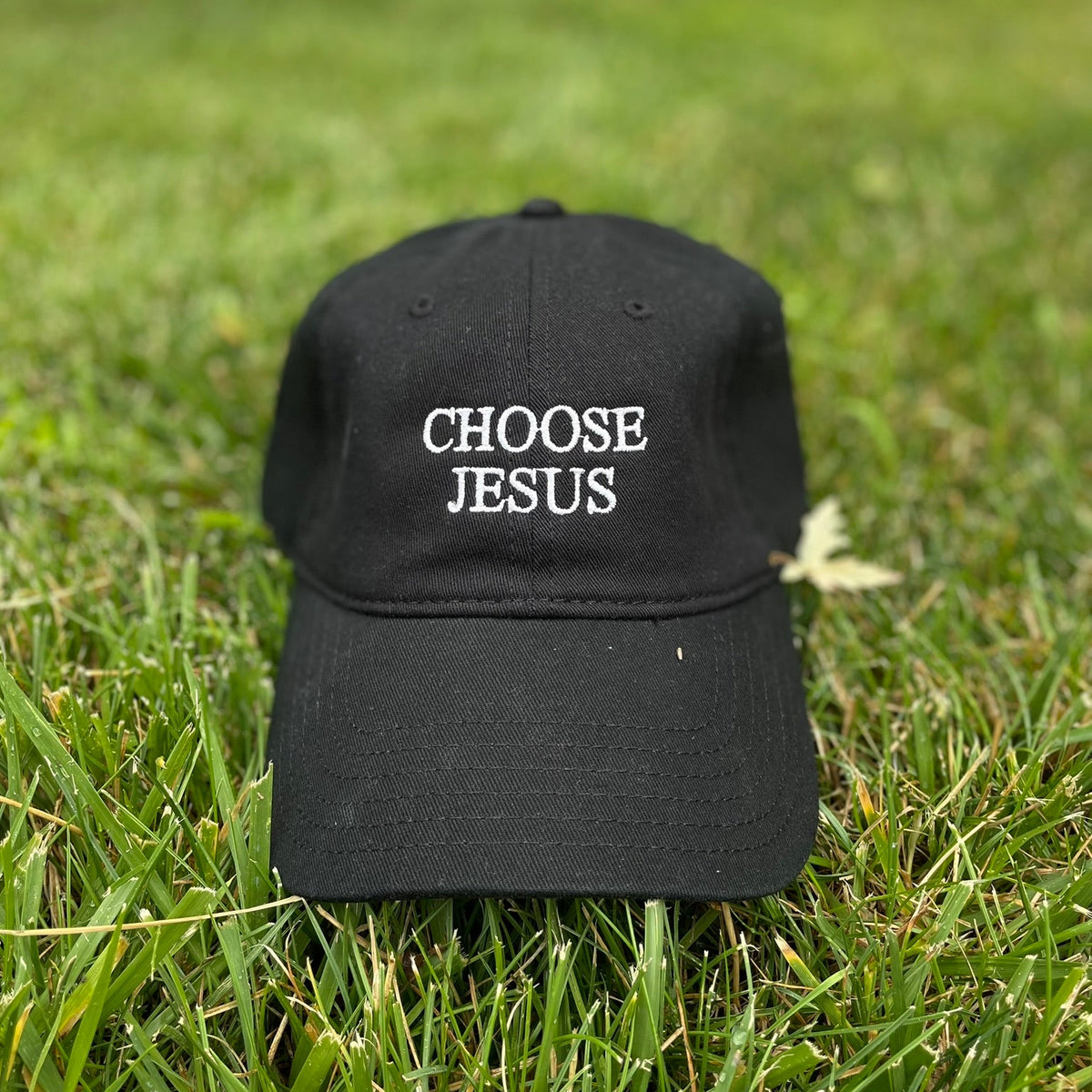 A black baseball cap with white embroidered text reading 'CHOOSE JESUS' sits on green grass outdoors.