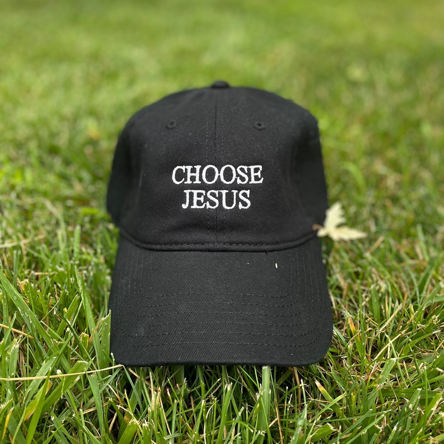 A black baseball cap with white embroidered text reading 'CHOOSE JESUS' sits on green grass outdoors.