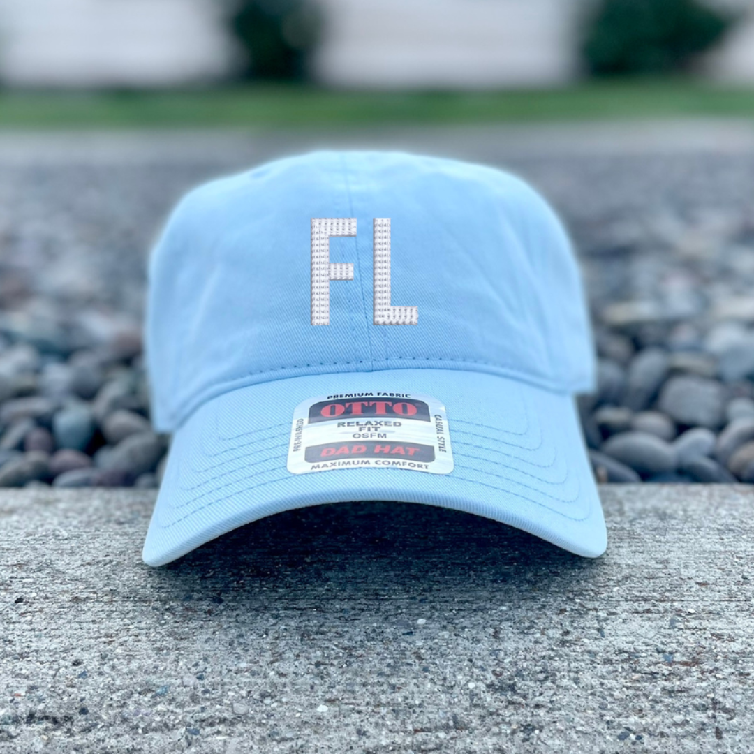 A light blue baseball cap with 'FL' embroidered on the front in a rhinestone pattern, resting on a concrete surface with gravel and a blurred house in the background.
