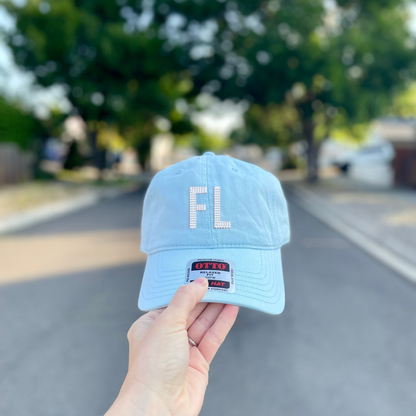 A light blue baseball cap held up by a hand against a blurred outdoor street background. The cap features a white rhinestone 'FL' logo on the front and a small rectangular patch on the brim.