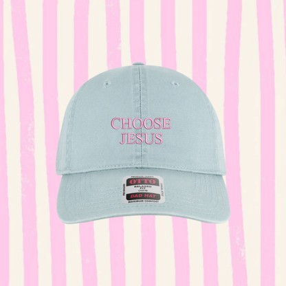 A light blue baseball cap with 'CHOOSE JESUS' embroidered in pink on the front, set against a pink and cream vertical striped background.