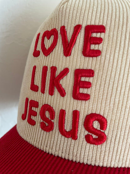 A close-up of a corduroy trucker hat with a cream crown and red brim, featuring red embroidered text that reads 'LOVE LIKE JESUS' on the front panel.