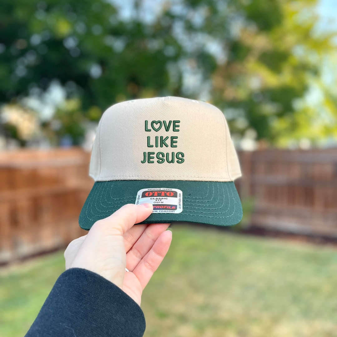 A hand holding a two-tone snapback cap outdoors with a blurred backyard background. The cap has a beige crown with dark green embroidered text reading 'LOVE LIKE JESUS' and a dark green visor.